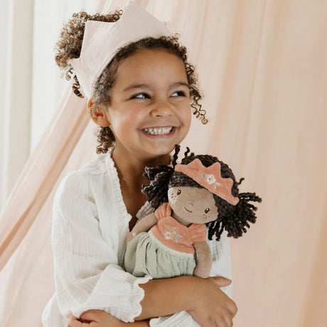 A smiling young child with curly hair holding a little dutch doll with dark curly hair, a peach crown, and a pale green dress