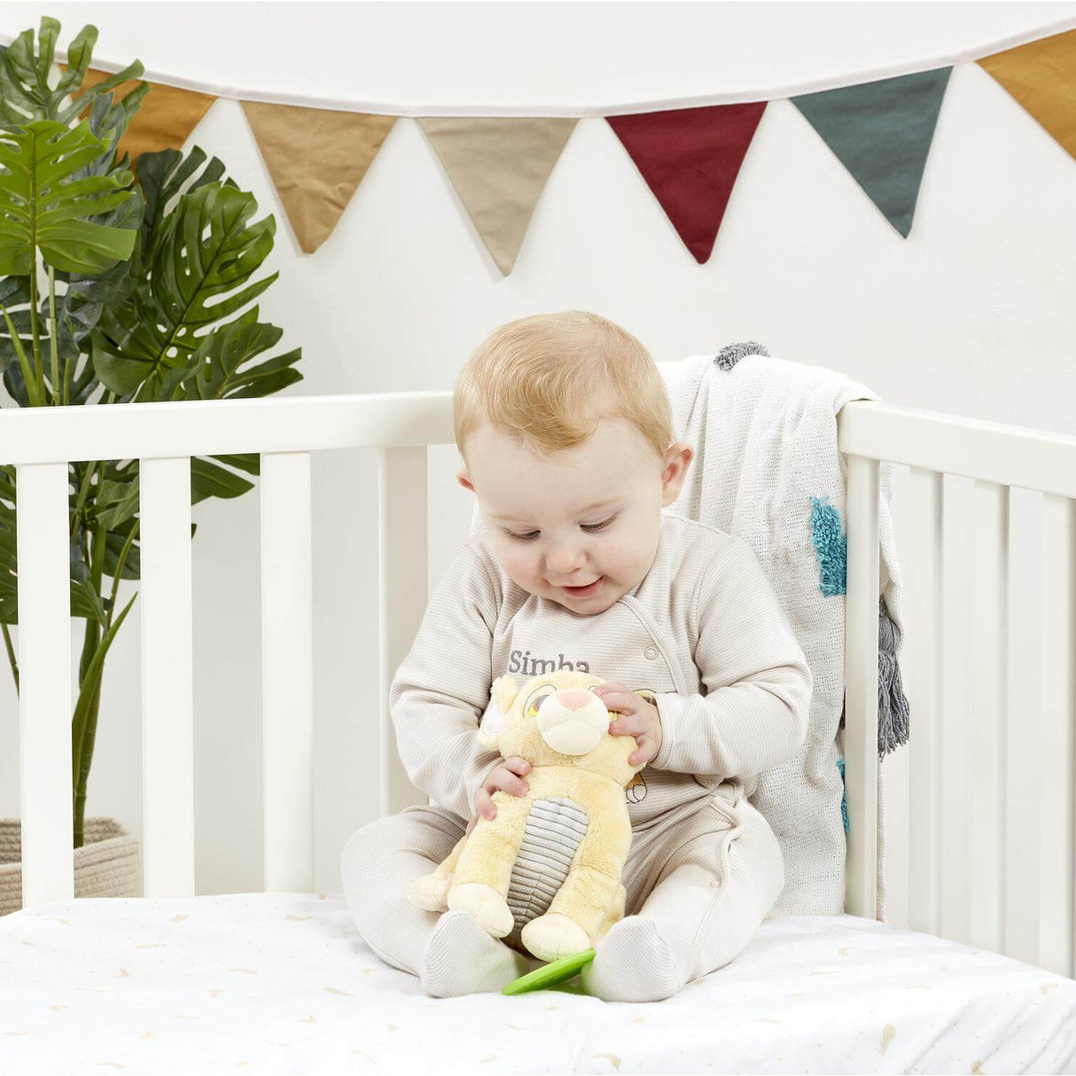 Baby sitting in a crib holding a Plush baby toy of a lion cub Simba from Disney's 'The Lion King' with green teether leaf attached with a colourful banner in the background