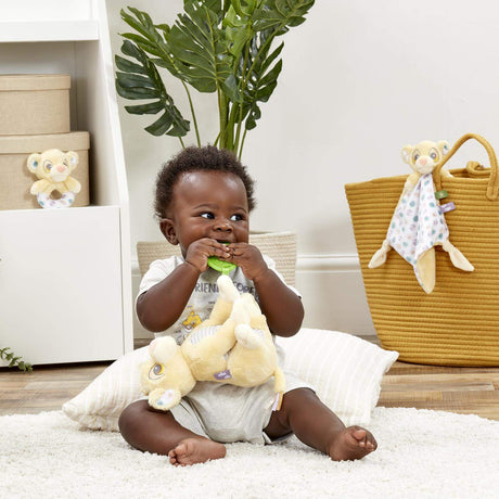 Baby sitting in a crib holding a Plush baby toy of a lion cub Simba from Disney's 'The Lion King' with green teether leaf attached with a colourful banner in the background