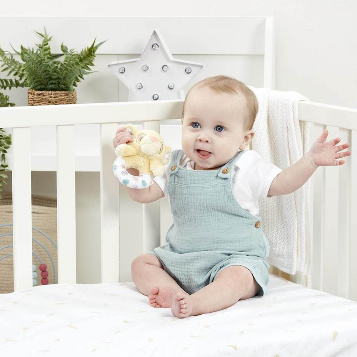 Baby sitting in a crib holding a Disney Lion King ring rattle, wearing a light blue romper and white shirt.