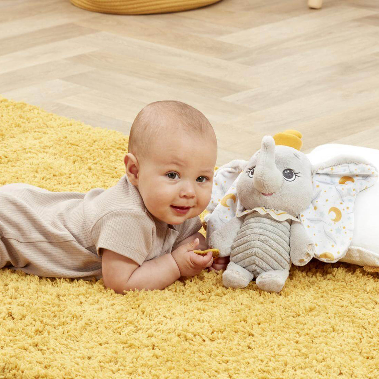Baby lying on a yellow rug next to the Disney Dumbo My First Soft Toy, holding the yellow star teether and smiling.