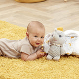 Baby lying on a yellow rug next to the Disney Dumbo My First Soft Toy, holding the yellow star teether and smiling.