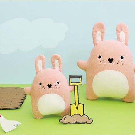Two pink plush bunnies with a shovel and sandcastle on a green surface with a light blue background.