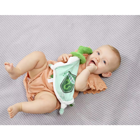 Baby lying on a textured surface with a plush Slytherin comfort blanket with a green and white color scheme, featuring a snake design, Slytherin crest, and embroidered stars.