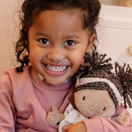 A smiling young girl with curly dark hair holds a plush doll with braided dark hair and a floral headband.