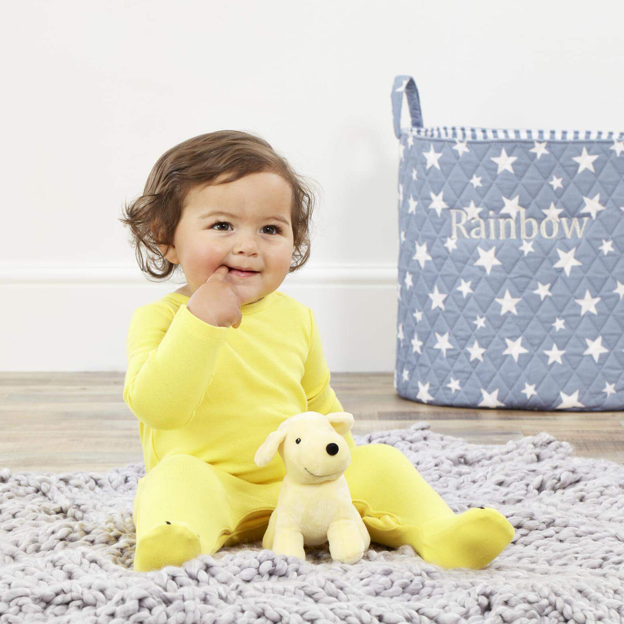 Child in yellow outfit with a yellow plush Spot the Dog soft toyon a textured rug, next to a star-patterned storage bag.