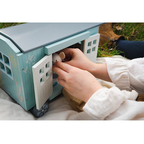 Small toy house with a hand opening a door, set outdoors on grass.