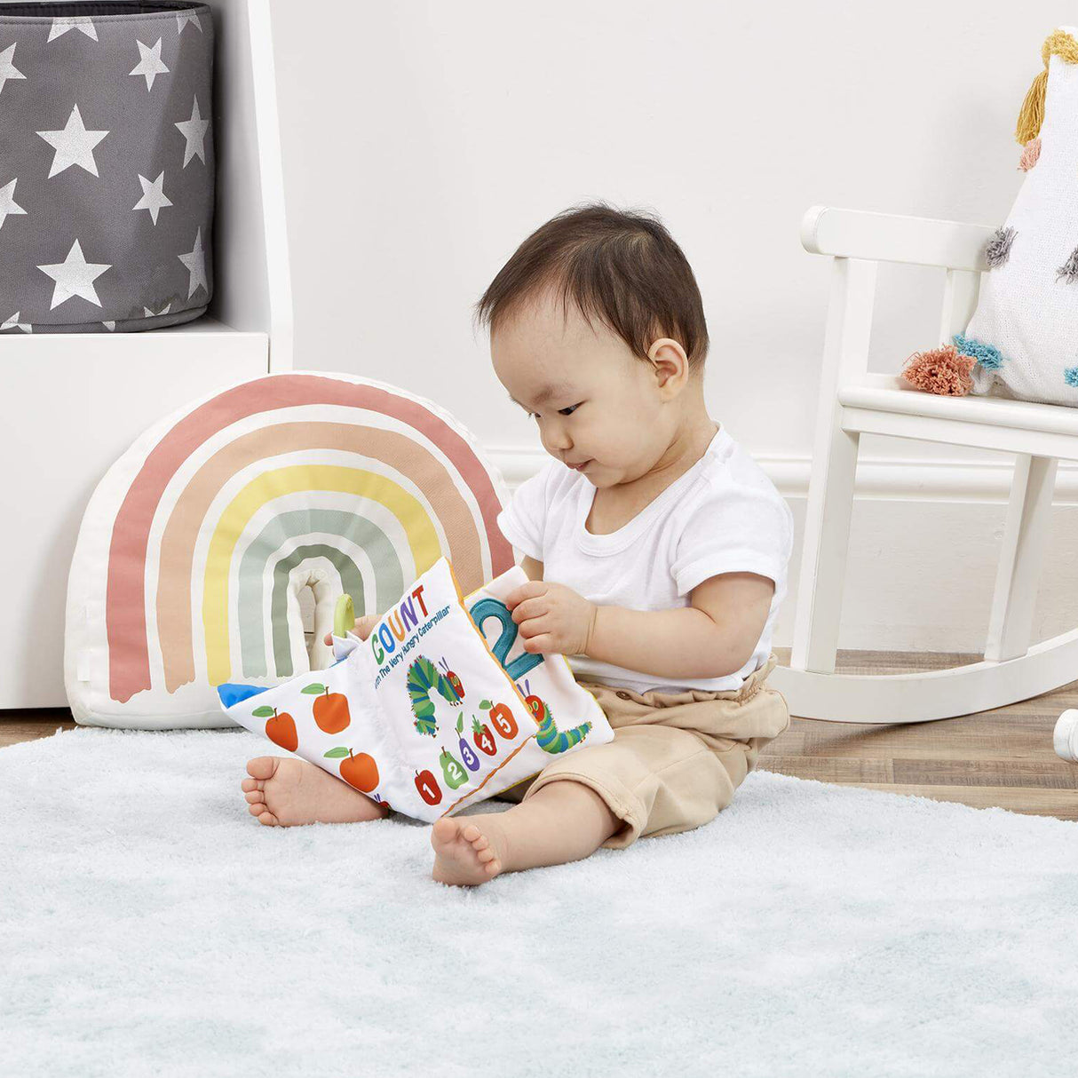 Baby sitting on a rug holding a soft baby book with a rainbow pillow in the background