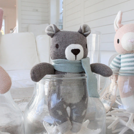 Knitted teddy bear with a light blue scarf  with a blue scarf inside a glass jar, surrounded by other toys.