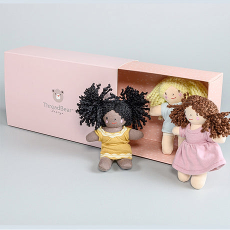 Three small rag dolls with different hairstyles and outfits in front of a pink 'ThreadBear Design' box.