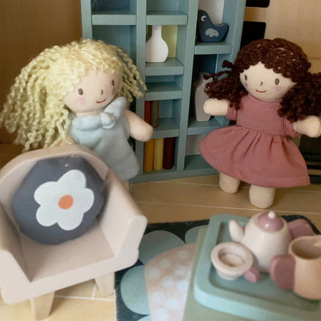 Two dolls with blonde and brown hair, a chair with a flower cushion, and a small table with a teapot set on a wooden floor.