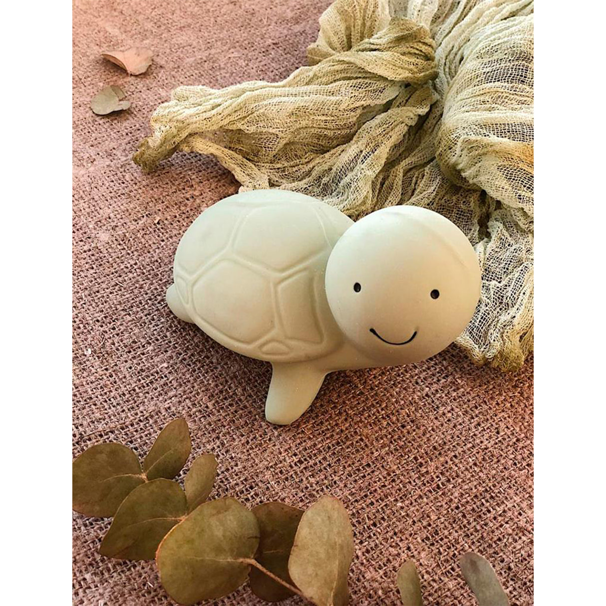 Green rubber rattle turtle-shaped bath toy with a smiley face on a textured pink surface with eucalyptus leaves.