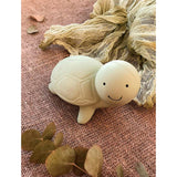 Green rubber rattle turtle-shaped bath toy with a smiley face on a textured pink surface with eucalyptus leaves.