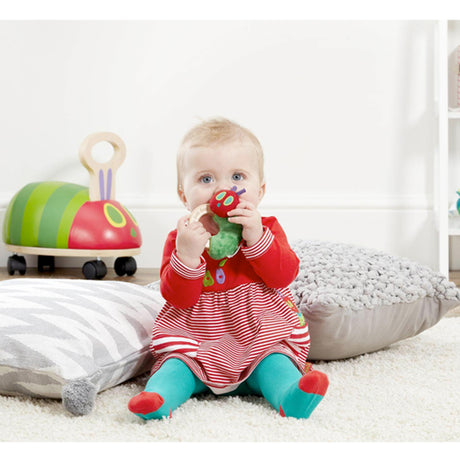 Baby in a red dress playing with a toy in a room with a white curtain and striped cushion.
