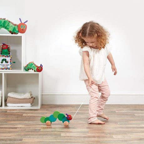 Child playing with a toy caterpillar on a wooden floor with shelves in the background.