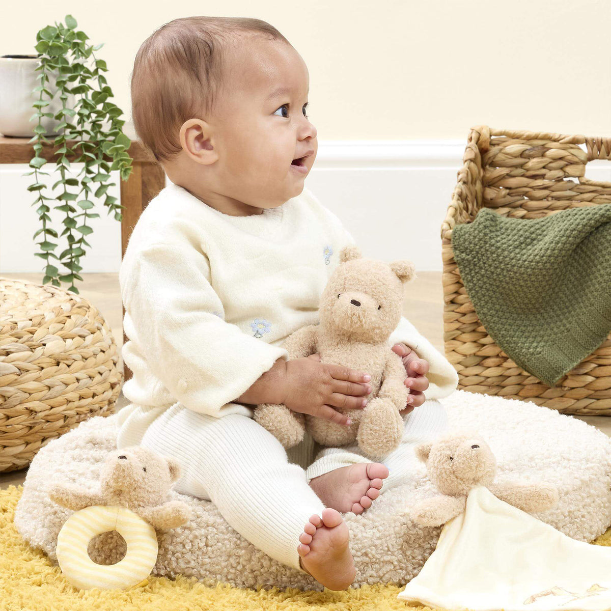 A baby sitting in a nursery setting, holding a small beige Pooh bear