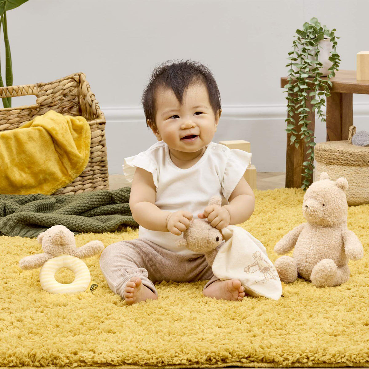 A baby sitting on a yellow shag rug holding a small pooh comforter, surrounded by plush toys and baby accessories in a styled nursery setting.
