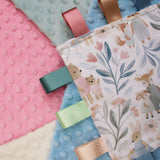 Close-up of a baby sensory taggy blanket with satin ribbon, behind is arranged pink, blue, green and cream minky dot fabric
