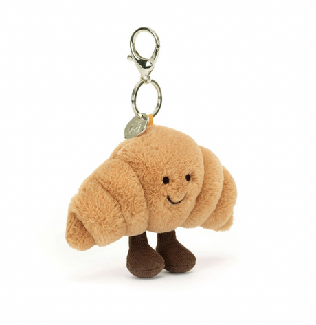 A fluffy golden croissant bag charm with chocolate cord boots and a smiling face, attached to a silver chain.