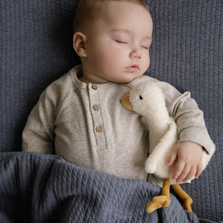 Baby cuddling a goose soft toy with a cream body and wings and a orange beak and  feet.